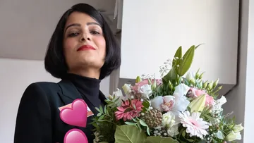 Person hält Blumenstrauß drinnen, schaut selbstbewusst.