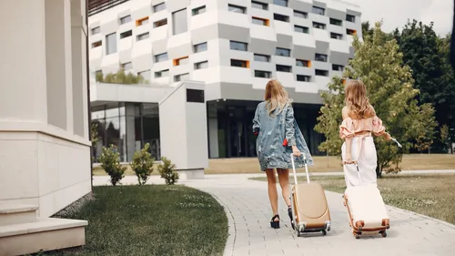 Two women with luggage walking towards building.