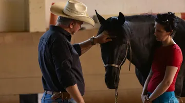 Man in cowboy hat gently touching horse indoors.