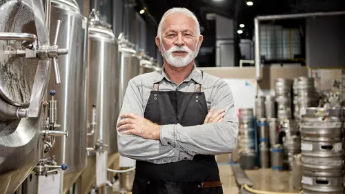 Man with apron stands in brewery with tanks.