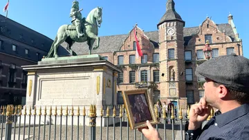 Man holding portrait by equestrian statue, historic building.