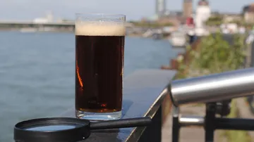 Glass of dark beer on railing, riverside view.