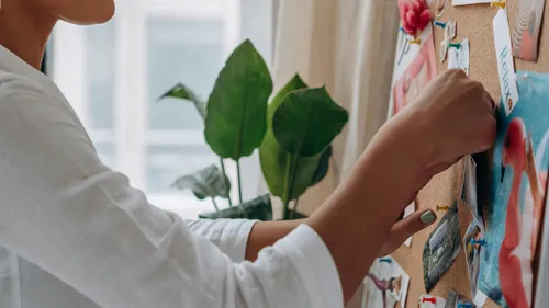 Person arranging photos on corkboard at window.