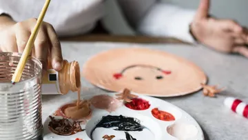 Hand pours paint onto palette near painted face.