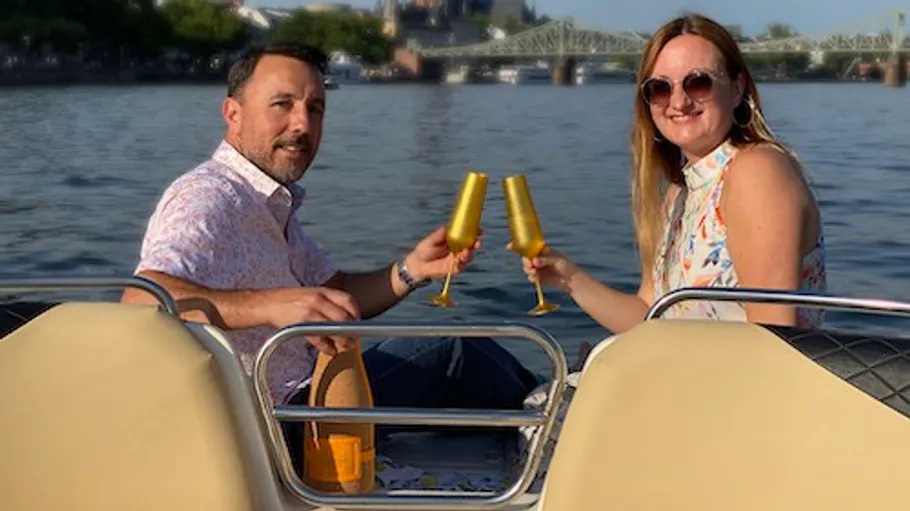 Paar stößt auf einem Boot an, Fluss im Hintergrund.