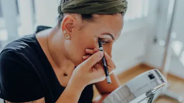Person applying eyeliner, seated by mirror.