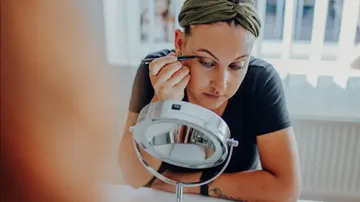 Woman applying eyeliner using mirror indoors.