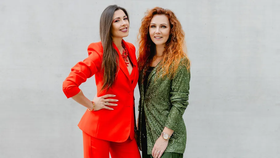 Zwei Frauen posieren in stilvollen Outfits, weißer Hintergrund.