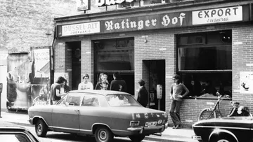 Menschen unterhalten sich vor Ratinger Hof Bar, Auto parkt.