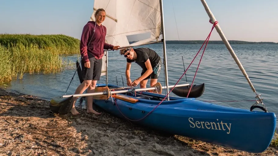 Two people preparing sailboat on lake shore.