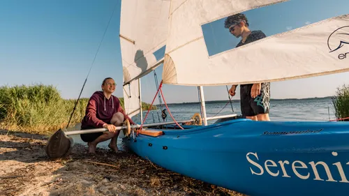 Two people with sailboat by a lake.
