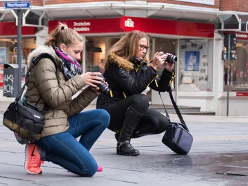 Two women take pictures kneeling in a town square.