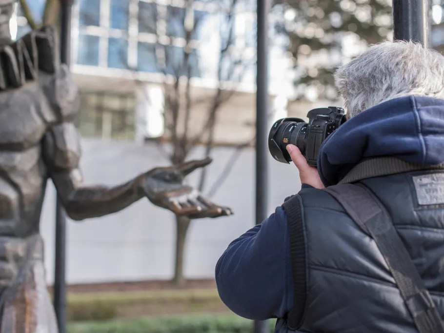 Mann fotografiert Statue in urbanem Umfeld mit Bäumen.