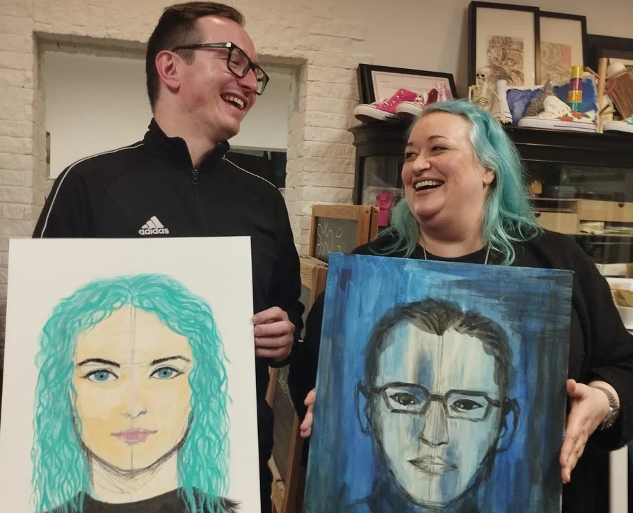 Two people smiling, holding painted portraits.