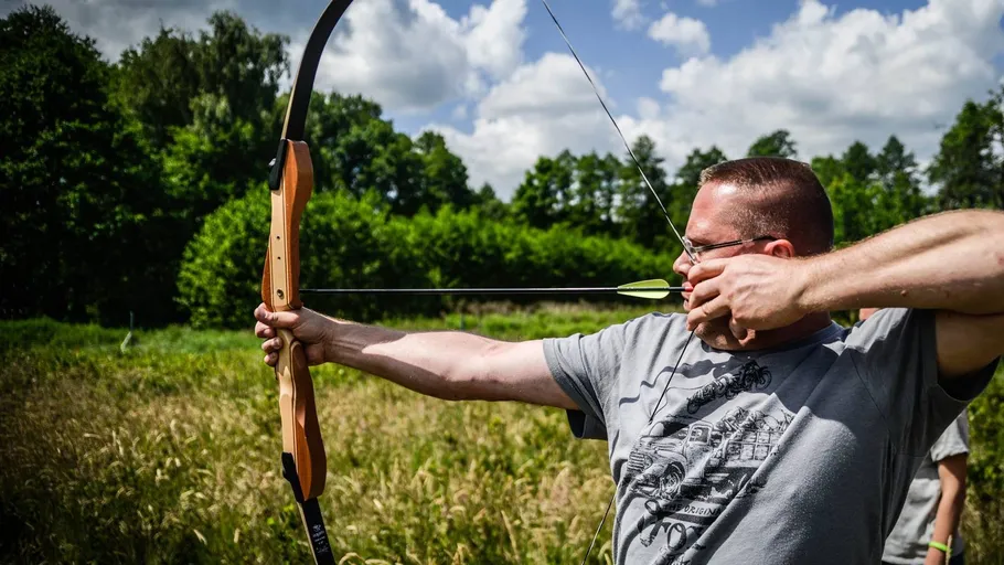 Mann zielt mit Bogen im Freien unter Bäumen.