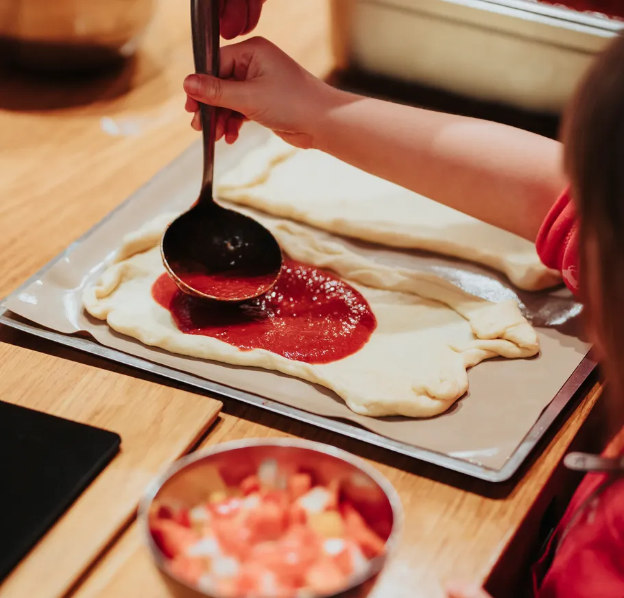 Child spooning sauce on dough in kitchen.