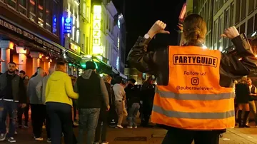 Person trägt 'Partyguide'-Weste in belebter Nachtstraße.