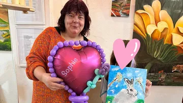 Frau hält Herzballon neben Kind mit Gemälde.