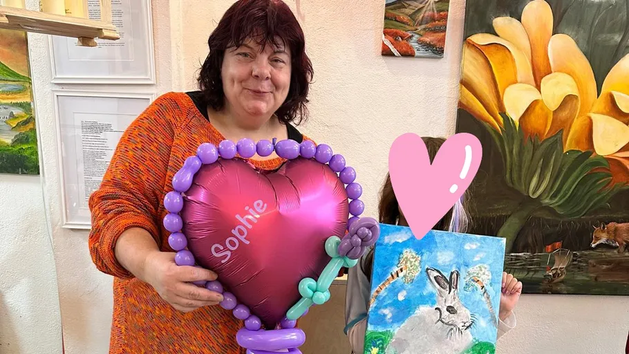 Woman holds heart balloon beside child with painting.