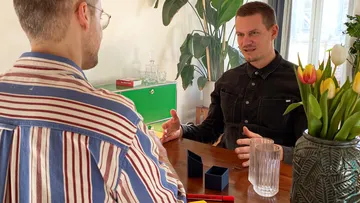 Zwei Männer sprechen an einem Holztisch.