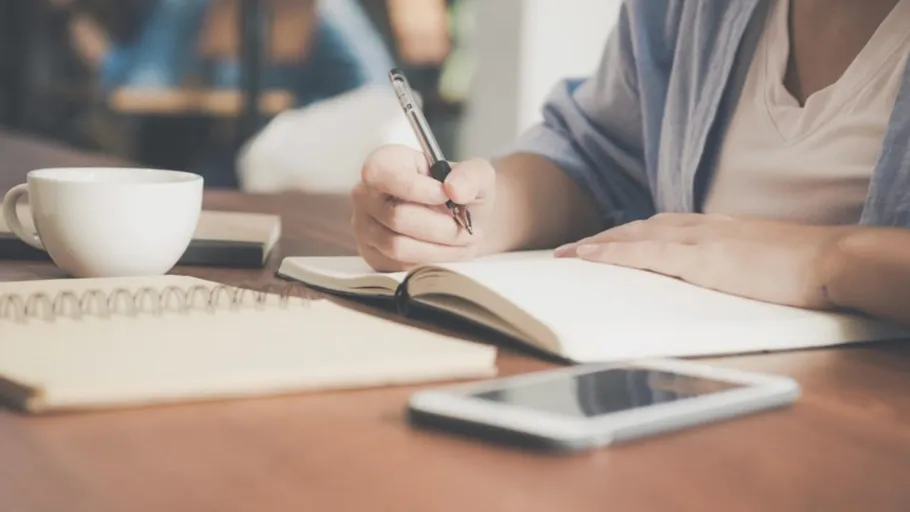 Person writing in notebook at wooden table.