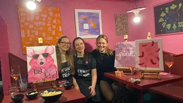 Three women smiling in a pink art studio.