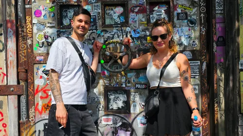 Two people posing near sticker-covered wall.