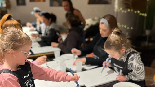 Kinder und Erwachsene malen an Tischen drinnen.