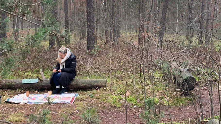 Person sitting on log in forest with picnic setup.