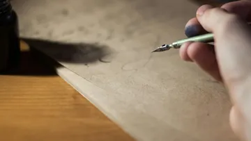 Hand writes with quill on paper, desk background.