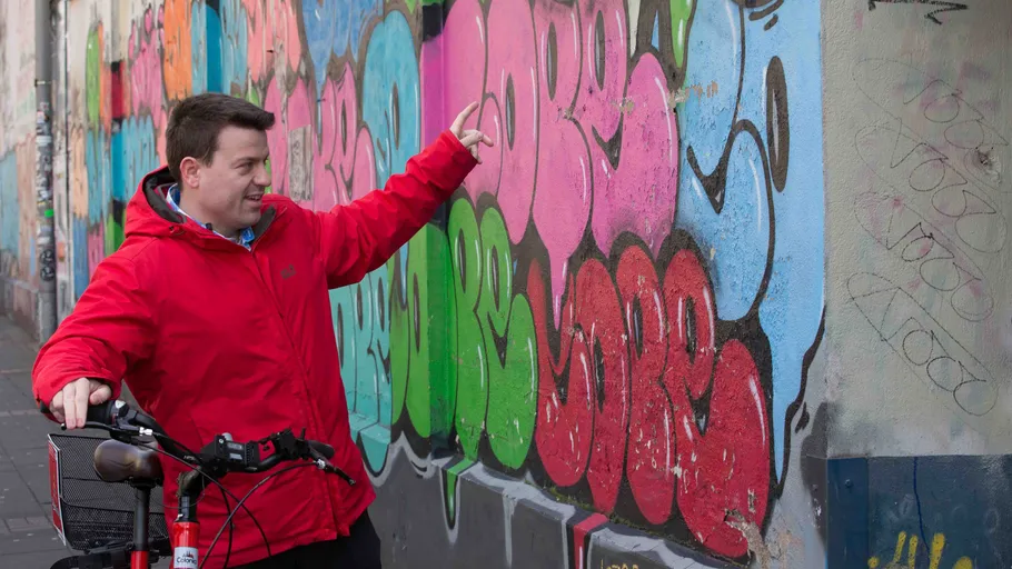 Man in red jacket points at colorful graffiti wall.