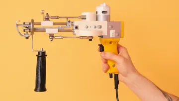 Hand hält Tufting-Pistole vor orangem Hintergrund.
