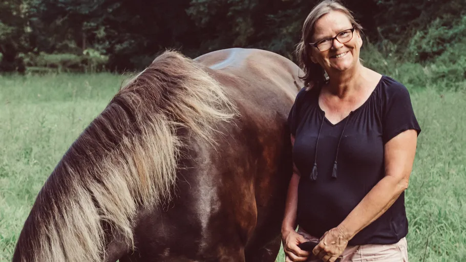 Frau lächelt neben grasendem Pferd auf Wiese.
