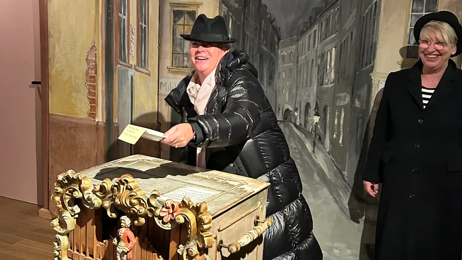Woman in hat plays street organ indoors.
