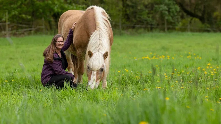 Frau kniet neben weidendem Pferd auf Wiese.