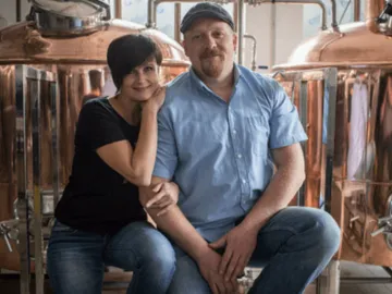 Two people pose in front of large copper kettles.