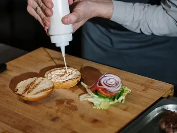Hand spritzt Sauce auf ein Brötchen, um Sandwich zu belegen.