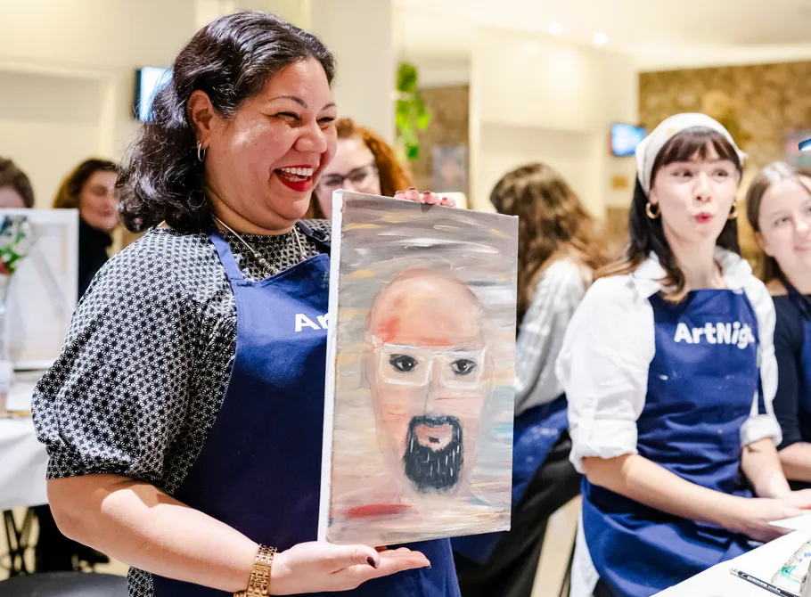 Smiling woman holds painting, people in background.
