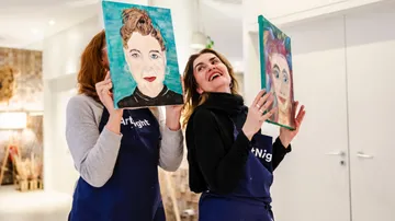 Two women hold painted portraits indoors.