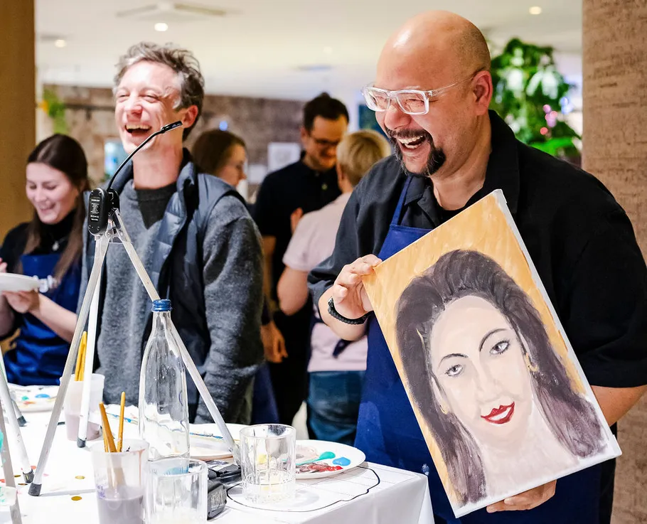 Two men laughing, displaying a painted portrait.