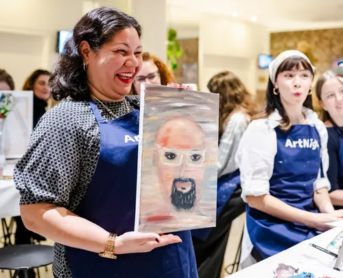 Woman displays painting, surrounded by group indoors.