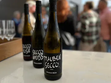 A bottle of grape secco is standing on a table with people in the background.