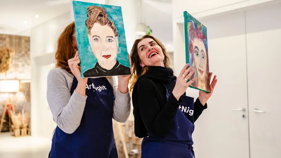 Two women holding paintings, smiling indoors.