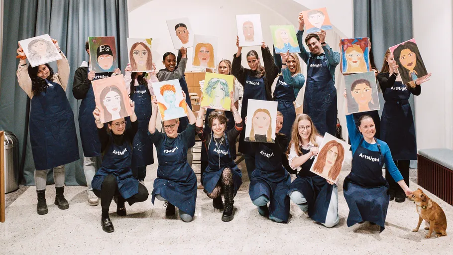 Group holding paintings, wearing aprons, smiling indoors.