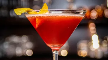 Red cocktail with orange garnish, bar background.