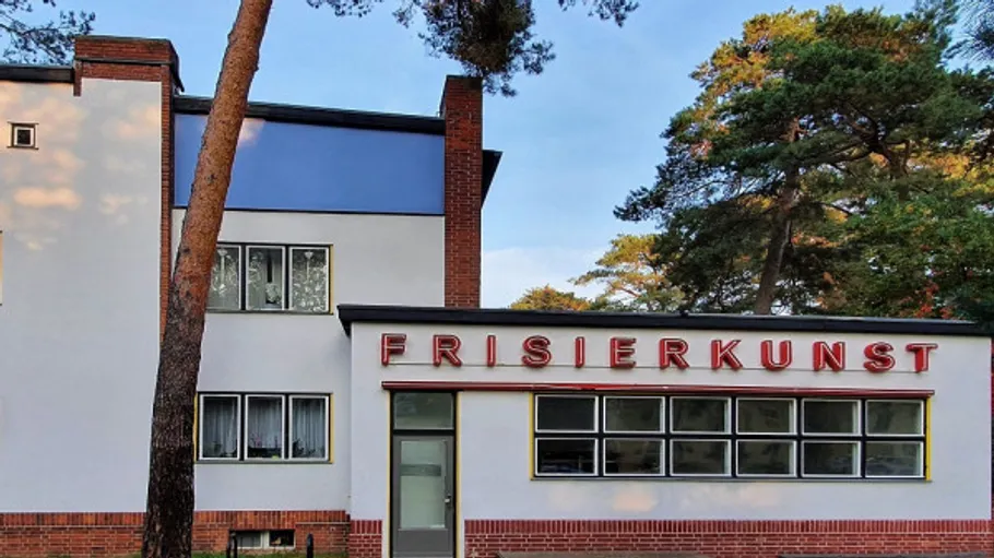 Gebäude mit 'Frisierkunst'-Schild, umgeben von Bäumen.