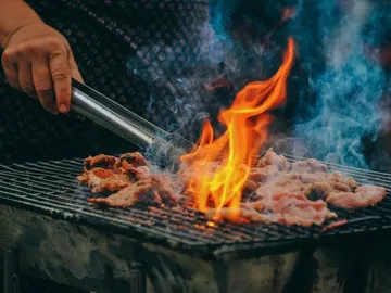 Meat is grilled over burning coals surrounded by smoke.