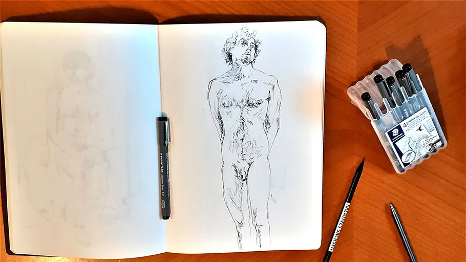 Sketchbook with human figure drawing, pens nearby.