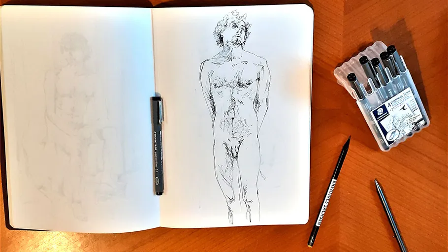 Sketchbook with human figure drawing, pens nearby.