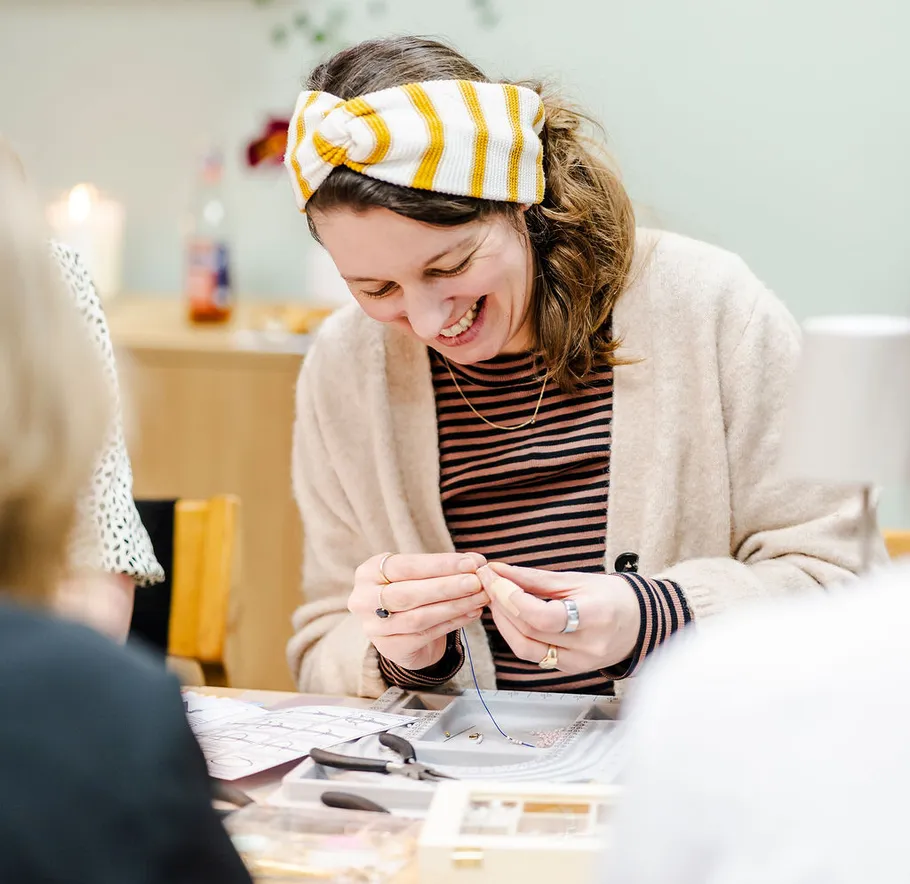 Frau bastelt Schmuck an einem Tisch, lächelnd.